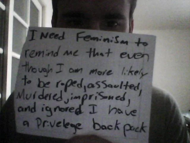 Who Needs Feminism — Person holding handwritten sign expressing support for feminism to address systemic dangers including sexual assault and murder.