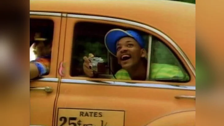 Will Smith With a Camera — Will Smith leans out of an orange taxi window wearing a blue cap and holding a camera, laughing.