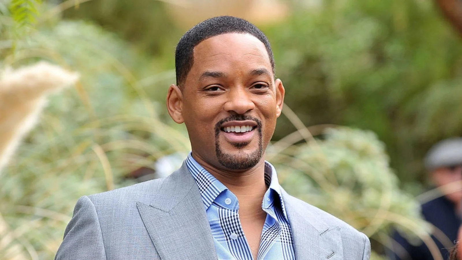 Actor Will Smith smiling in a grey suit jacket against a blurred leafy background