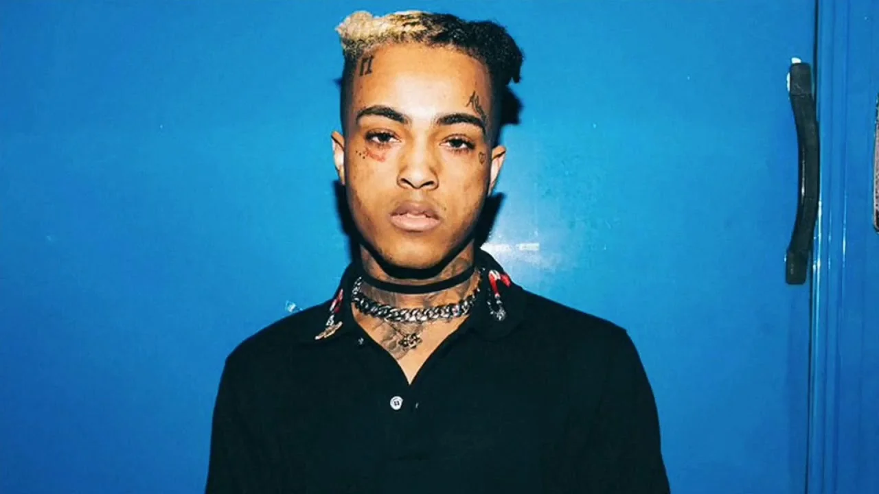 Portrait of XXXTentacion in a black polo against a blue wall