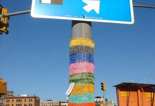 Yarn Bombing — Street sign pole wrapped in alternating bands of yellow, gray, blue, and pink knitted yarn.