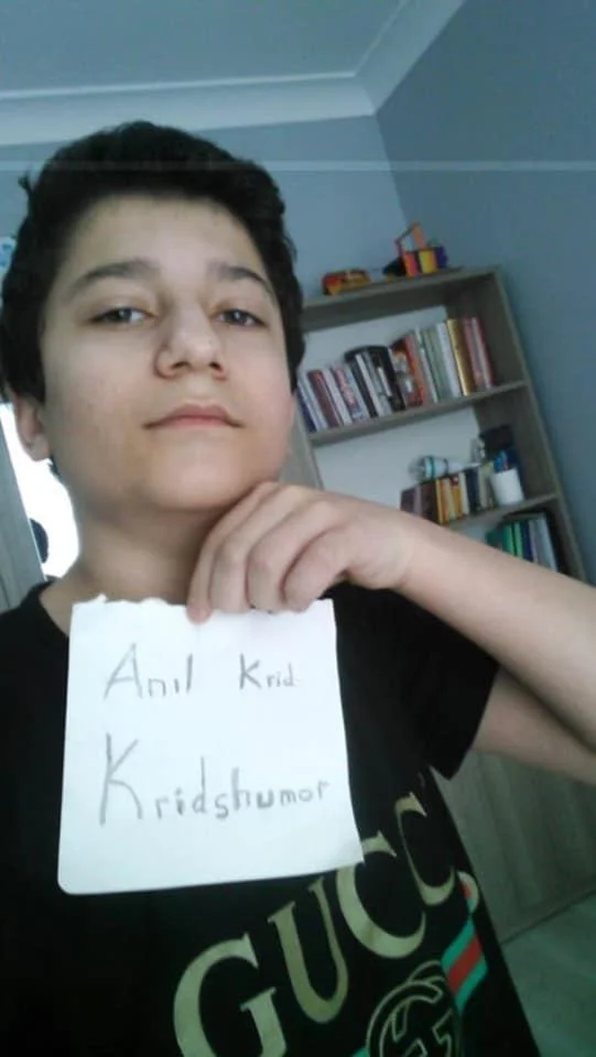 Yotube — Teen holding handwritten sign reading Anil Krid Kridshumor in front of a bookshelf, wearing a Gucci shirt