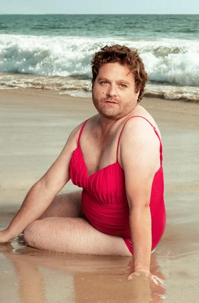 Zach Galifianakis in a red one-piece swimsuit sitting on a sandy beach